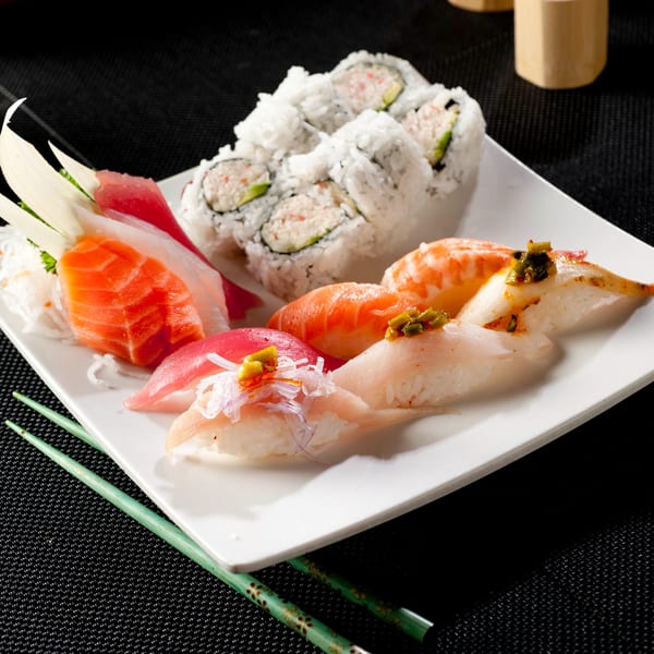 A white plate of Yoshi Japanese Cuisine sushi, sashimi, and California rolls with green chopsticks.