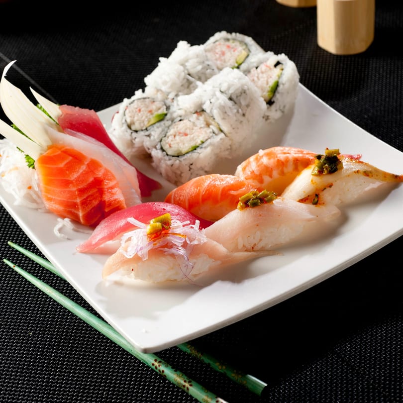 A white plate of Yoshi Japanese Cuisine sushi, sashimi, and California rolls with green chopsticks.